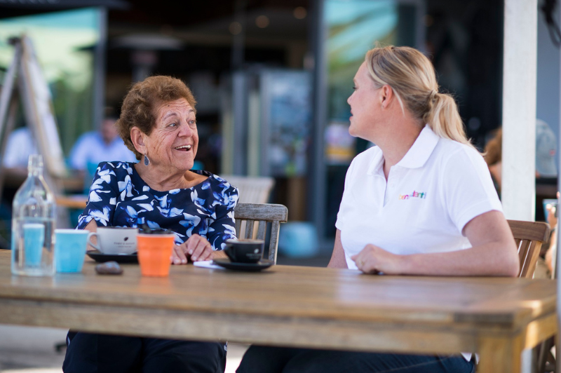 An aged care client smiling with her support worker at a cafe table